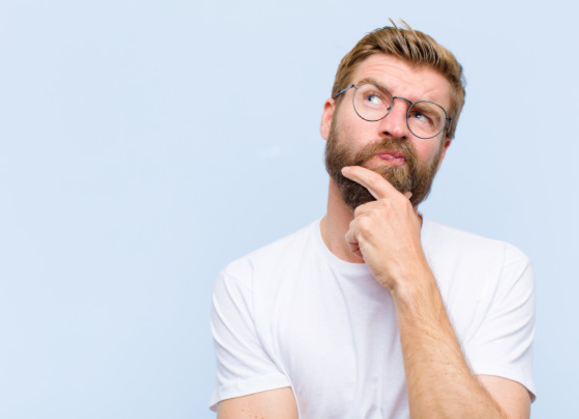 homem de óculos e camiseta branca pensativo em frente a um fundo azul claro