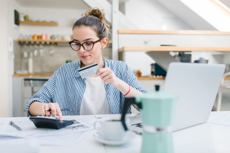 mulher com o cartão de crédito nas mãos e uma calculadora para realizar as contas da sua fatura