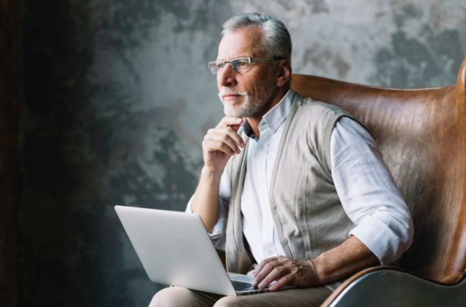 homem idoso de óculos e suéter nos ombros sentado em uma poltrona de couro usando um laptop