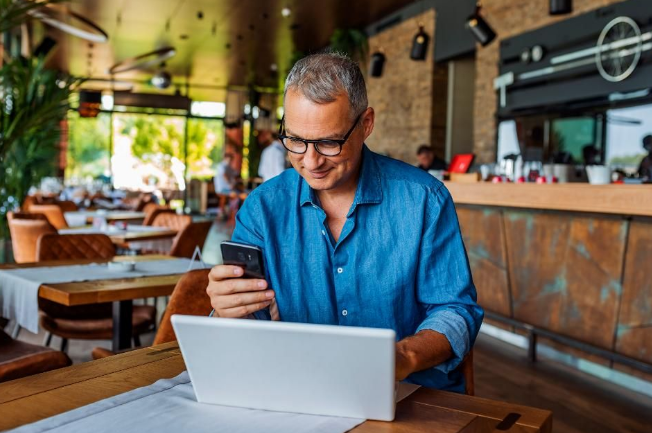 homem de camisa azul mexendo em sei celular em frente ao seu laptop