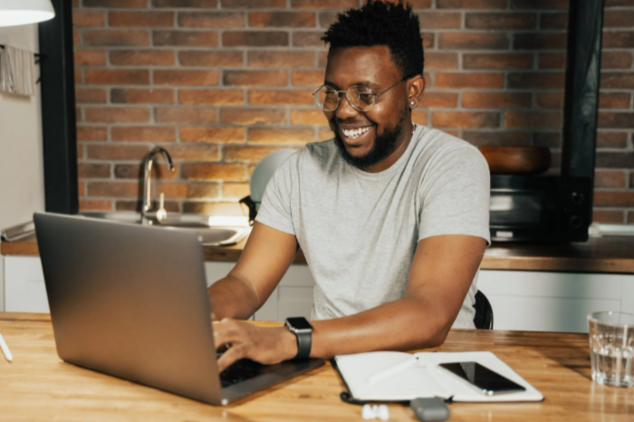 homem sorrindo sentado à mesa frente ao computador e ao lado de um caderno e um celular