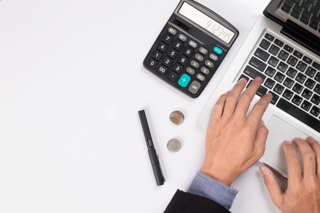 imagem de uma mesa com um laptop sendo operado por um homem de terno e camisa roxa ao lado de uma calculadora, uma caneta e um par de moedas