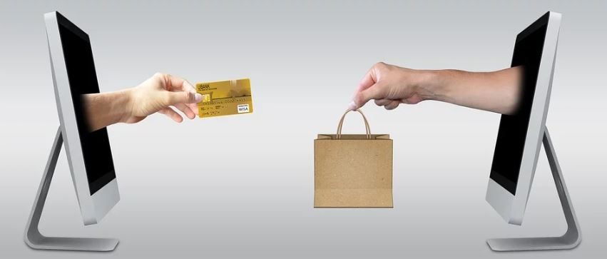 mão saindo de um monitor entregando uma sacola de compras para mão saindo de outro monitor entregando cartão de crédito.