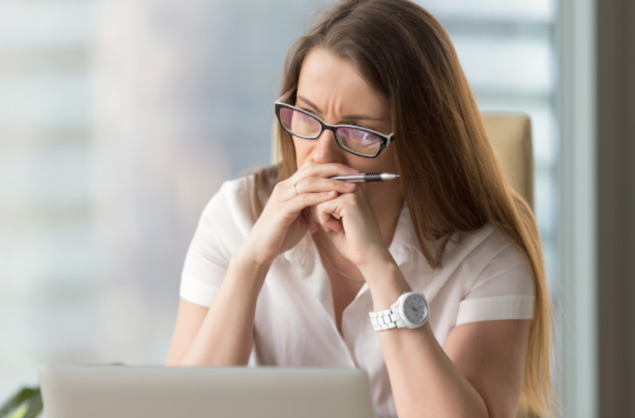 mulher de óculos usando roupas brancas sentadas em frente a laptop segurando uma caneta com semblante preocupado